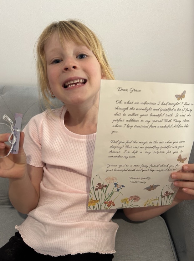 A child grinning with a gap in her teeth, holding the tooth fairy letter and a rolled note tied with ribbon.